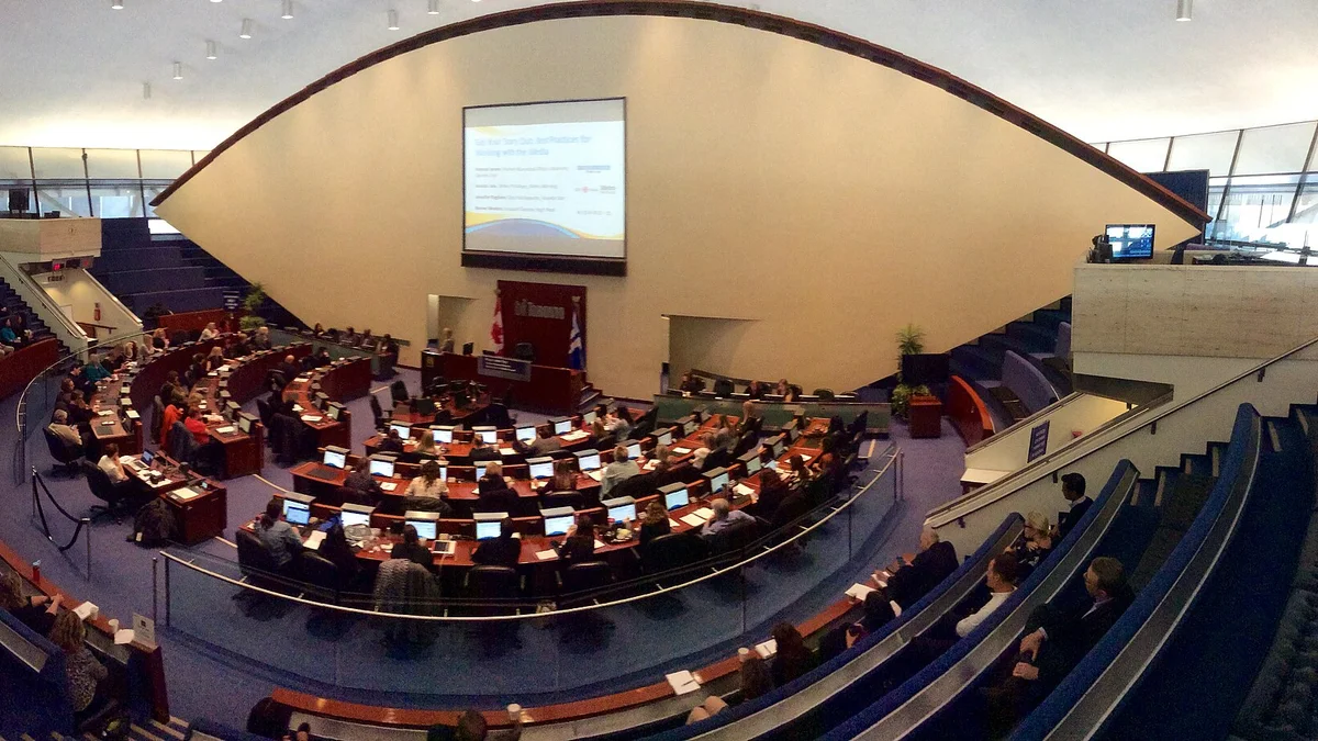 A city council chamber in session, with officials seated at curved desks before an audience