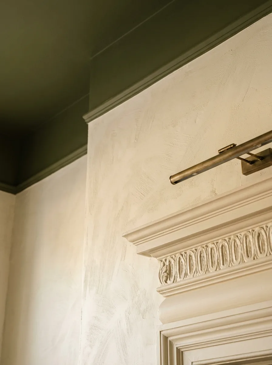 A limewashed plaster wall meeting a deep-olive color-drenched ceiling with a slim picture light and carved cornice
