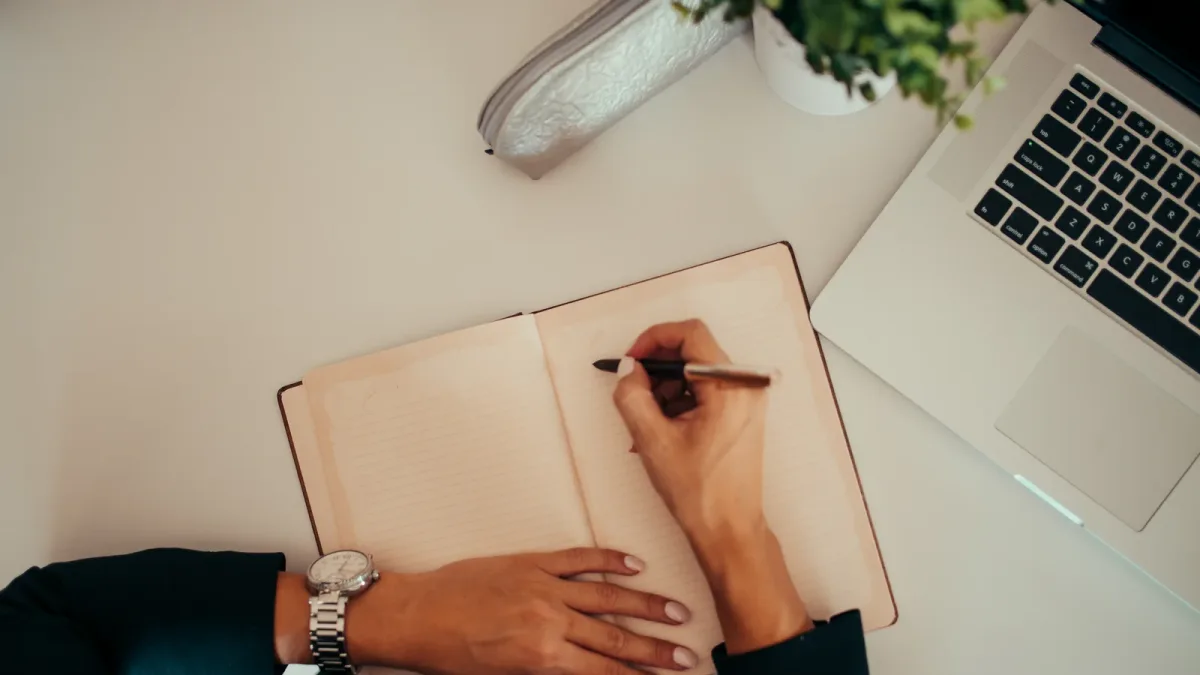 A person writing in a notebook beside a laptop at a modern workspace