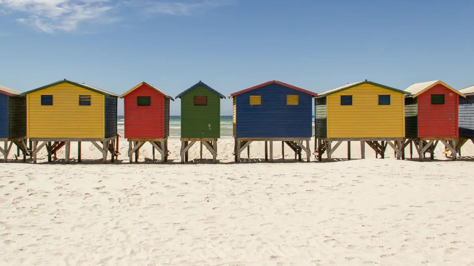 A row of brightly colored beach houses standing on the sand at Muizenberg Beach