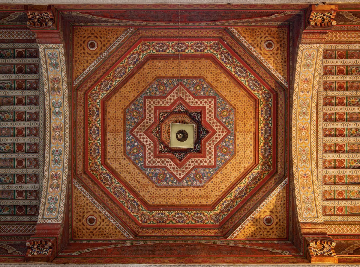 A carved and painted wooden ceiling in red, gold and green geometric patterns with a lantern hanging from the center
