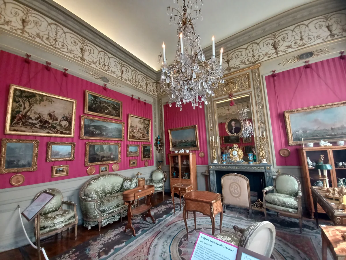 A salon with raspberry pink walls, gilded paintings, a crystal chandelier, upholstered chairs and a patterned carpet