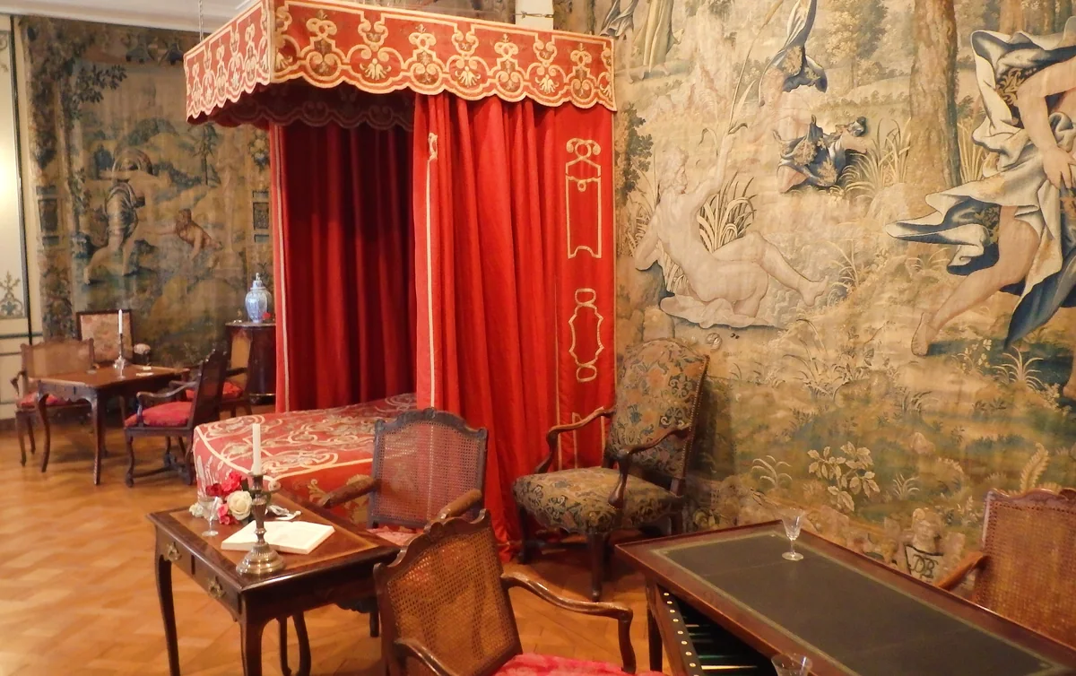 A canopy bed hung in red wool in a room lined with large tapestries, with antique chairs and a writing table