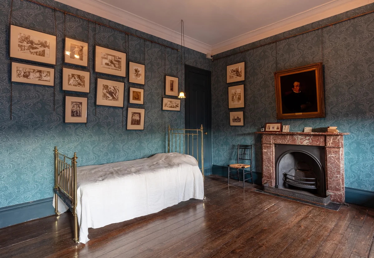 A plain bedroom with blue grey walls, a narrow brass bed with white covers, a fireplace and small framed drawings