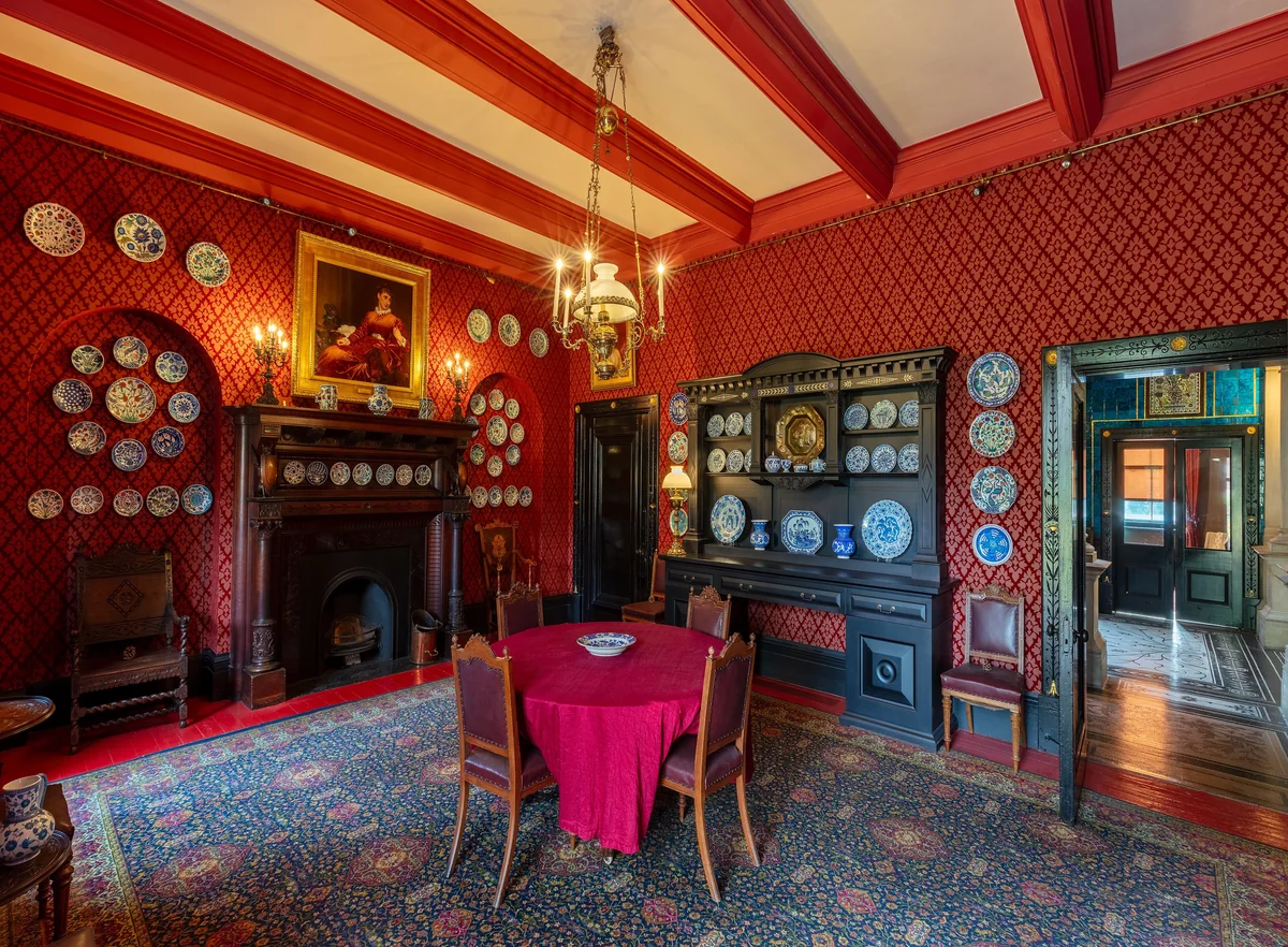 A dining room with deep red patterned walls, blue and white plates hung on the walls, a round table with a red cloth, a chandelier and a patterned rug