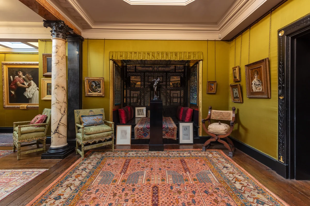 An ochre yellow drawing room with marble columns, framed paintings, upholstered chairs and a large patterned carpet
