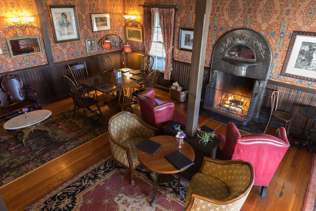 A hotel lounge with patterned wallpaper, red velvet chairs, a fireplace, framed pictures and Persian rugs