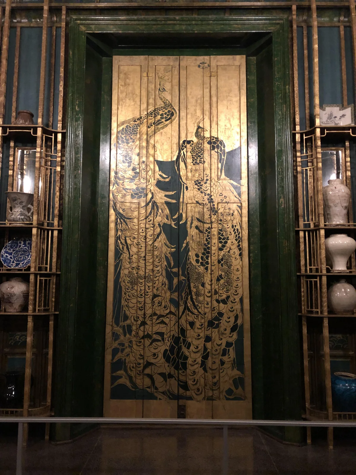 Gold painted peacocks on blue green lacquered shutters inside the Peacock Room, with shelves of blue and white porcelain