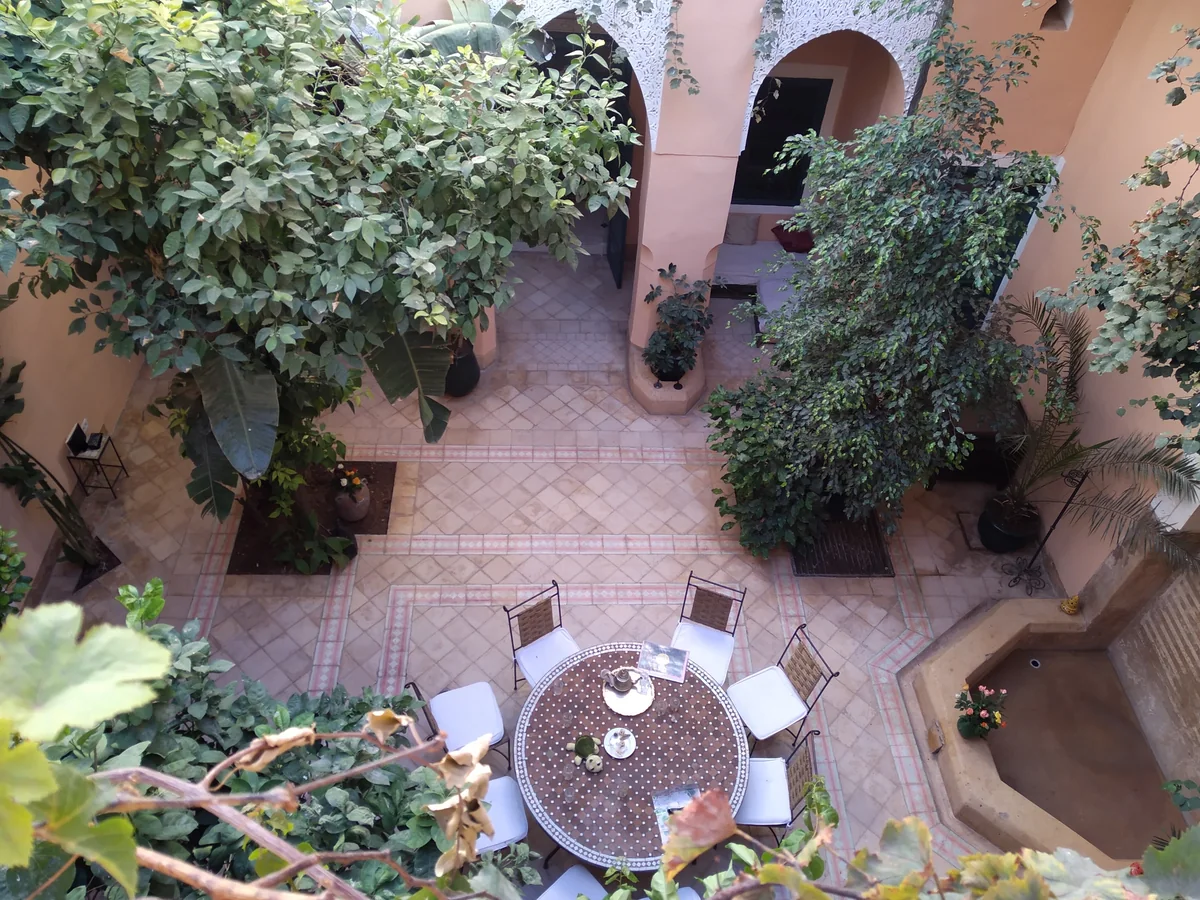 A riad courtyard seen from above with a tiled floor, a round table and chairs, and dense green plants on every side