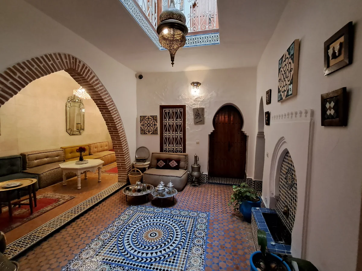 A riad lounge with low sofas, a hanging metal lantern, a geometric tiled floor, a blue patterned rug and an arched doorway