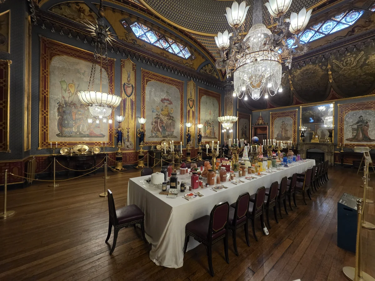A long banqueting room with a set table, painted wall panels, a painted ceiling and a large chandelier