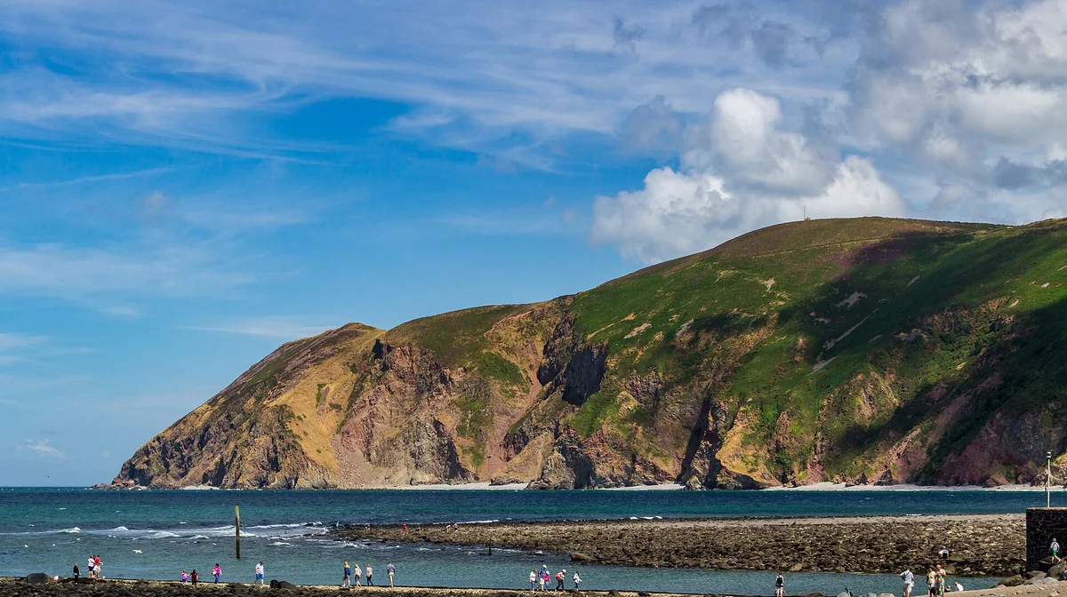 People walking along the rocky shore at Lynmouth on the north Devon coast, with a long bank of stones running into the sea and a steep headland of green slopes and red cliffs across the bay